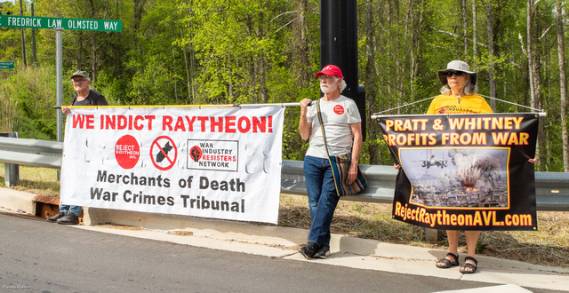 Protesters stand on the street outside the Pratt-Whitney plant with big banners, reading "We indict Raytheon! Merchants of Death War Crimes Tribunal" and "Pratt & Whitney Profits from War"